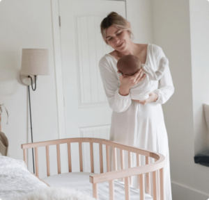 Mom holding baby by a bedside co sleeper after a successful baby shower | babybay bedside sleeper